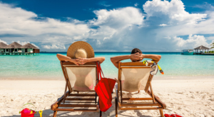 Couple in loungers on a tropical beach at Maldives looking out to villas on water and turquoise ocean.